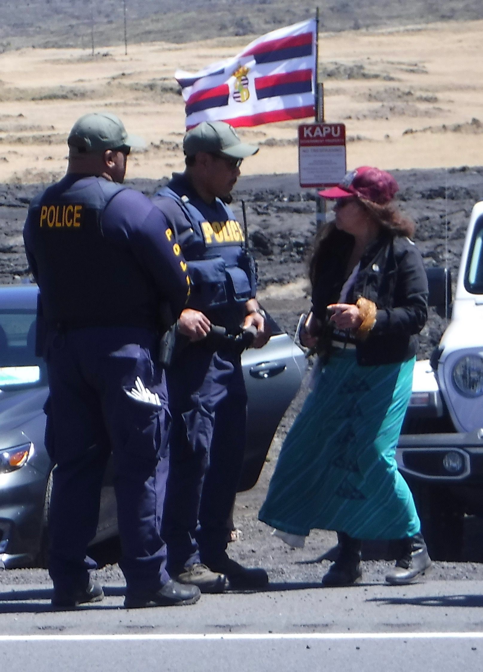 Indigenous woman being confronted by two police officers with cars and a flag in the background