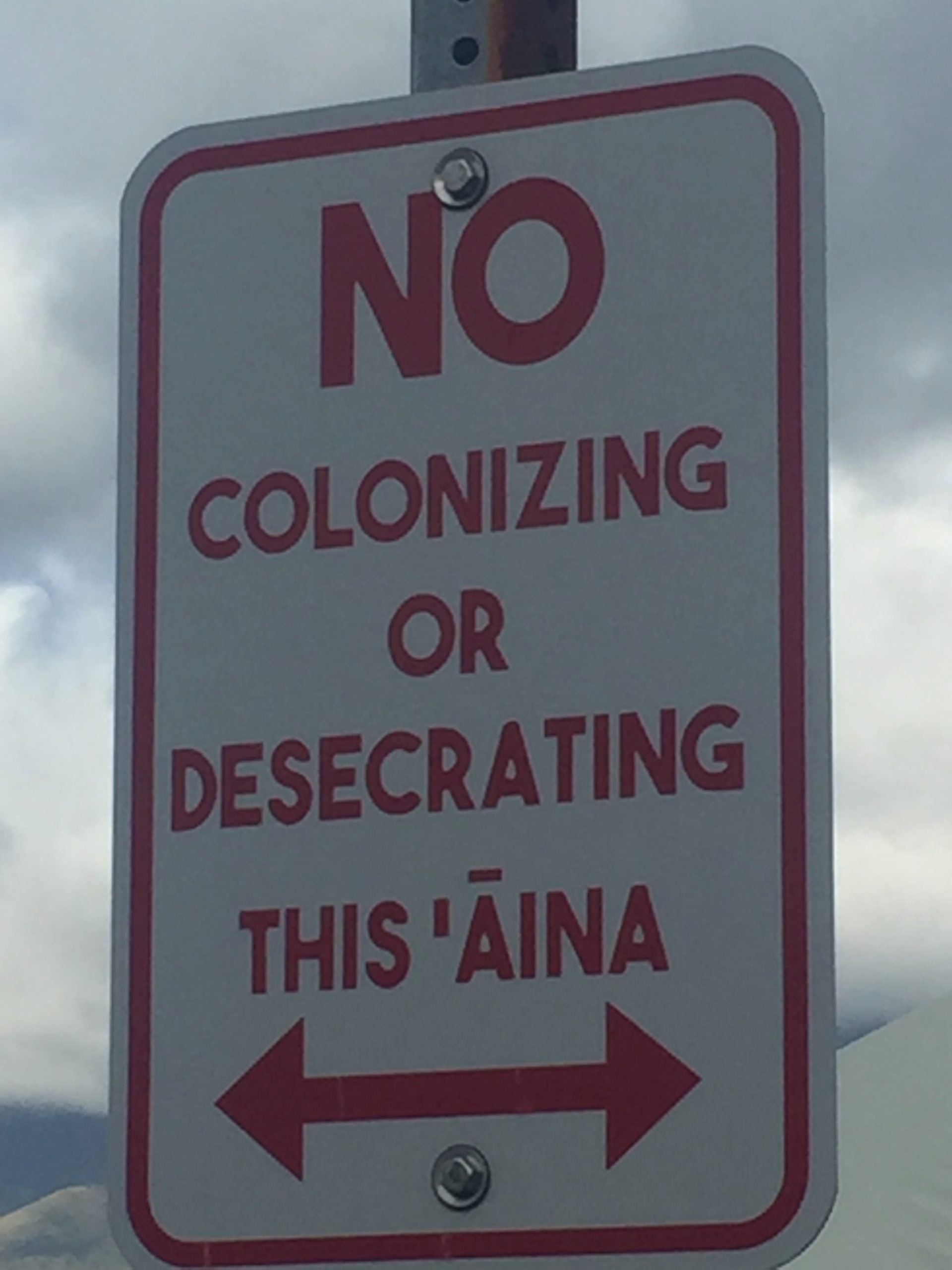 Close up of sign that reads "No colonizing or desecrating this aina (land/area) with double sided arrow